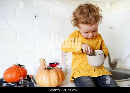 Cute little girl are sitting on the table near halloween pumpkins at home and eating tasty dessert Stock Photo