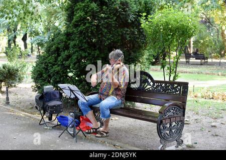 Old man sitting on a bench in the park and playing violin. Stock Photo