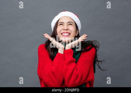 Surprised excited beautiful Asian woman in red Christmas sweater and hat smiling with open hands under chin studio shot on isolated gray background Stock Photo