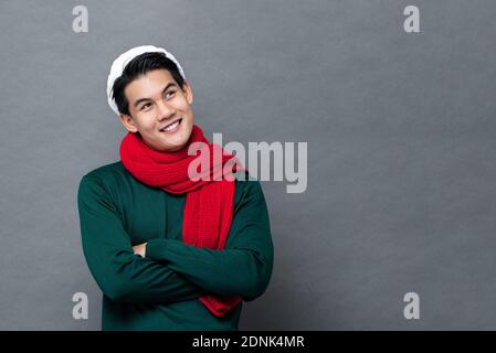 Young handsome Asian man in green Christmas sweater smiling and looking up at copy space with arms crossed isolated on gray background Stock Photo