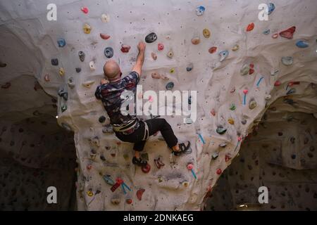 Active bald sporty man wearing protective face mask practicing rock climbing on artificial rock in a climbing wall. New normal in extreme sports. Stock Photo