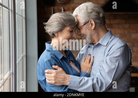 Loving elderly retired mature family couple cuddling near window. Stock Photo