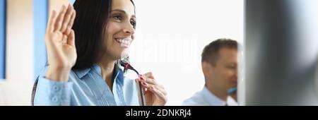 Businesswoman sits at computer with microphone smiling and raising her hand in a welcoming manner. Stock Photo