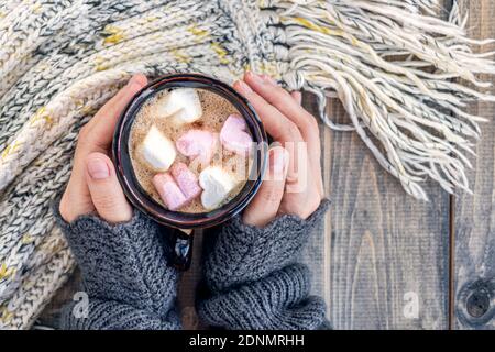 Female hands hold cup with a hot drink with marshmallows on of wooden table with warm knit woolen scarf. Christmas background - The concept of winter, warmth, holidays and events.Soft focus.Top view Stock Photo