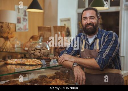 Handsome mature bearded male baker smiling to the camera proudly, working at his bakery shop, copy space. Friendly baker with manbun hairdo leaning on Stock Photo