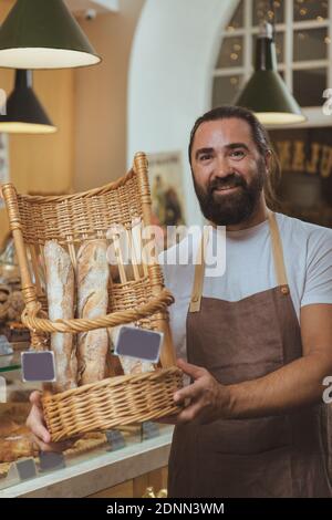 Vertical shot of a handsome cheerful baker offering you delicious fresh bread from the basket he is holding. Mature man working at his bakery, selling Stock Photo