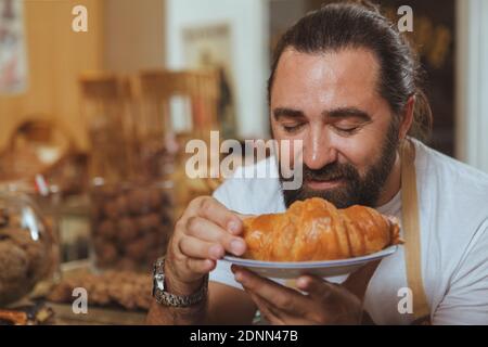 Close up portrait of a cheerful bearded male baker smelling delicious croissant, copy space. Handsome mature man working at the bakery, enjoying smell Stock Photo