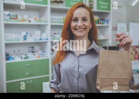 Lovely mature red haired woman smiling joyfully to the camera, holding paper shopping bag after buying medications at the drugstore. Happy female cust Stock Photo
