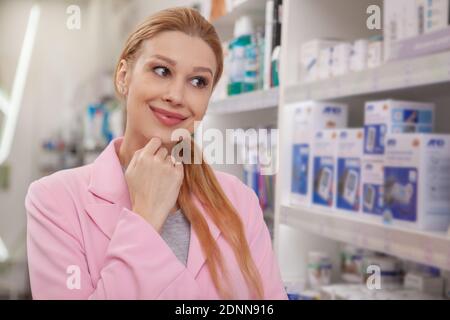Young lovely woman smiling joyfully, thinking which medicine to buy, shopping at local drugstore. Attractive female customer buying medication at phar Stock Photo