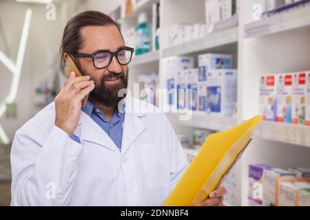 Handsome mature bearded pharmacist talking on the phone, selling medication online. CHeerful male chemist working at pharmacy, reading medical papers Stock Photo