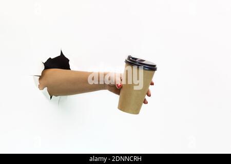 A woman's hand with red manicure through torn white paper holds a paper cup of coffee. Hand through torn white paper.Hand holding paper cup Stock Photo