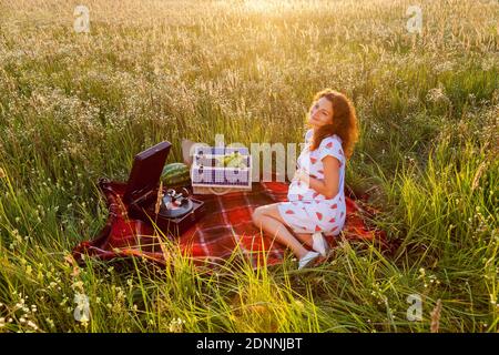 A pregnant woman is sitting on the red blanket near a gramophone and a basket with fruits on the wheat field on a sunny day. Stock Photo