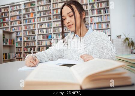 Young Asian female student writing in her notebook at the library, open book on foreground Stock Photo