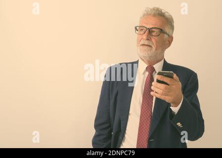 Studio shot of handsome senior bearded businessman thinking while holding mobile phone against white background Stock Photo