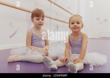 Two adorable cheerful little ballerinas rest on the floor at dance studio. Cute happy little girl in ballet outfit smiling joyfully, sitting with her Stock Photo