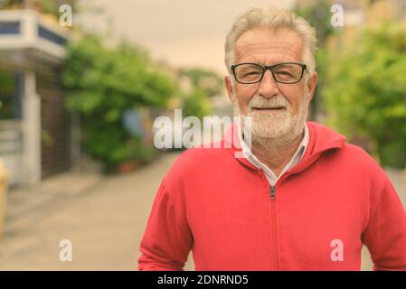 Happy handsome senior bearded man smiling while wearing eyeglasses and red jacket outdoors Stock Photo