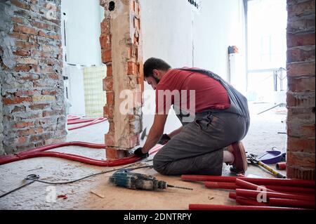 Man plumber in work overalls using hammer while laying pipes on the floor. Worker installing heating system while working on renovation of apartment. Concept of home renovation. Stock Photo