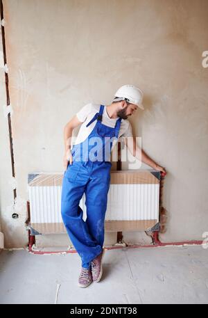 Full length of bearded young man plumber checking radiator while installing heating system in apartment. Male worker wearing blue work overalls and protective helmet, standing near heating battery. Stock Photo