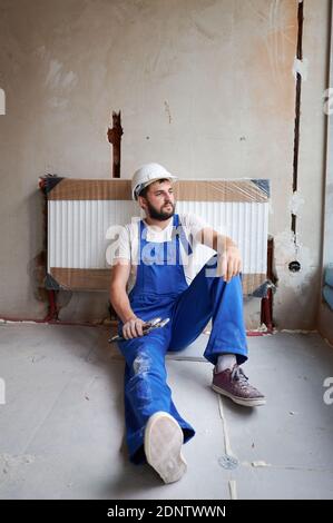 Young plumber wearing working uniform and helmet, sitting on the floor, looking away, holding wrench, resting on radiator after finishing installation works. Concept of heating system installation. Stock Photo