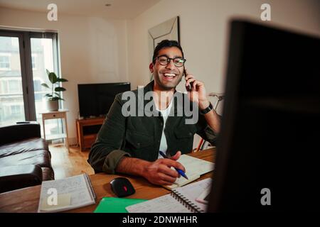 Happy mixed race business man chatting to family on smartphone while working from home office Stock Photo