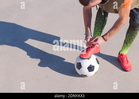 unrecognizable football player tying his boots with a foot on the ball in a concrete soccer field, concept of healthy lifestyle and urban sport in the Stock Photo