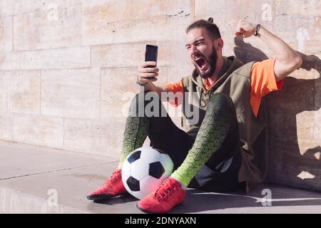 hipster football fan celebrates a victory when he is checking his smart phone, concept of technology and urban sport lifestyle in the city, copy space Stock Photo