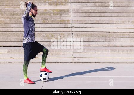 sportsman picking up his hair in a tail with a foot on his football ball in a concrete soccer court, concept of healthy lifestyle and urban sport in t Stock Photo