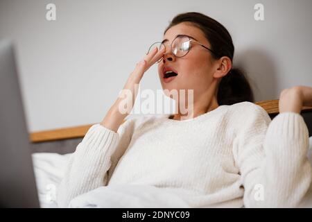 Exhausted young woman with laptop computer in bed working at home, yawning Stock Photo