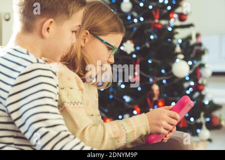 Happy teen siblings children or friends playing games with tablet on sofa near christmas tree at home Stock Photo