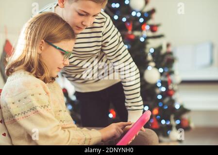 Happy teen siblings children or friends playing games with tablet on sofa near christmas tree at home Stock Photo