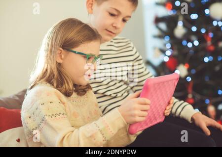 Happy teen siblings children or friends playing games with tablet on sofa near christmas tree at home Stock Photo