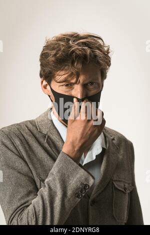 Close up portrait of young business man in a disposable facial mask in grey jacket and a modern reusable protective mask on his face fixing it Stock Photo