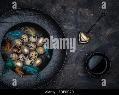 Easter concept. Quail eggs, golden colored eggs and colorful feathers in black bowl. Cup of black tea and tea strainer. Overhead view with copy space. Stock Photo