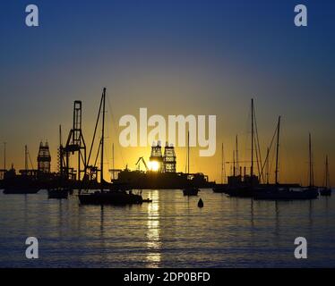 Sunrise from the port, backlit sailboats in the foreground, large ships and cranes in the background and clear blue sky Stock Photo