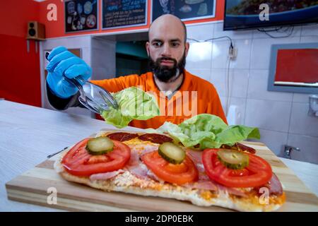 Chef preparing tasty fresh baguette submarine sandwich. Close up of hands in gloves holding tongs and arranging fresh lettuce over the baloney. Stock Photo