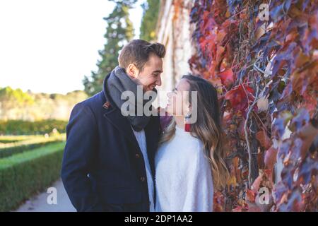 Young couple smile at each other beside the red leaf wall in the park on a sunny winter day Stock Photo