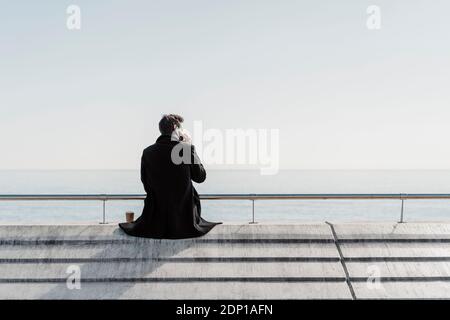 Businessman sitting on quay wall talking on cell phone Stock Photo