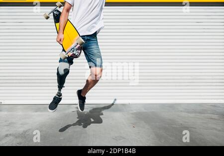 Young man with leg prosthesis holding skateboard and jumping Stock Photo