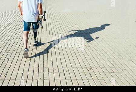Young man with leg prosthesis walking and holding skateboard Stock Photo
