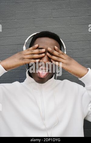 Portrait of young man listening music with headphones covering eyes while sticking out tongue Stock Photo