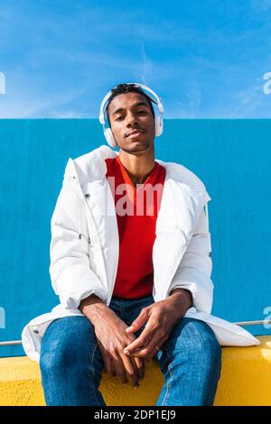 Young trendy man sitting on yellow wall, listening music Stock Photo