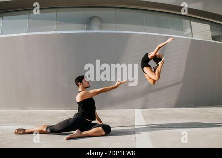 Female dancer doing acrobat while male gymnast practicing stag leap by gray wall Stock Photo