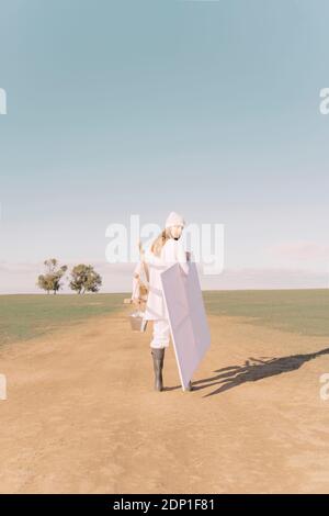 Young woman carrying easel and empty canvas on dry field Stock Photo