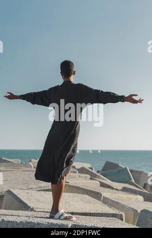 Young man wearing black kaftan on concrete blocks at the coast Stock Photo