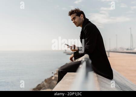 Businessman sitting on quay wall with cell phone Stock Photo