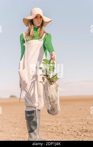 Young woman standing on stool on dry field, carrying plant in a net Stock Photo