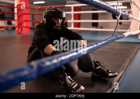 Exhausted woman in protective gear and sportswear sitting on floor and resting after boxing match in contemporary gym Stock Photo