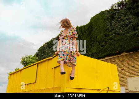 Woman in flower dress sitting on edge of yellow container Stock Photo