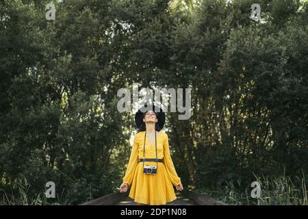Young woman wearing a black hat and yellow dress with an analog camera on wooden boardwalk Stock Photo