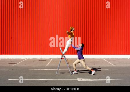 Young man and woman performing with a ladder in front of a red wall Stock Photo